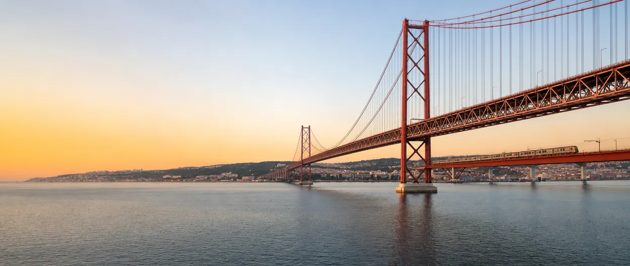 The red 25 de Abril suspension bridge over the Tagus at sunset, with a train crossing on the lower rail deck and Lisbon on the far bank